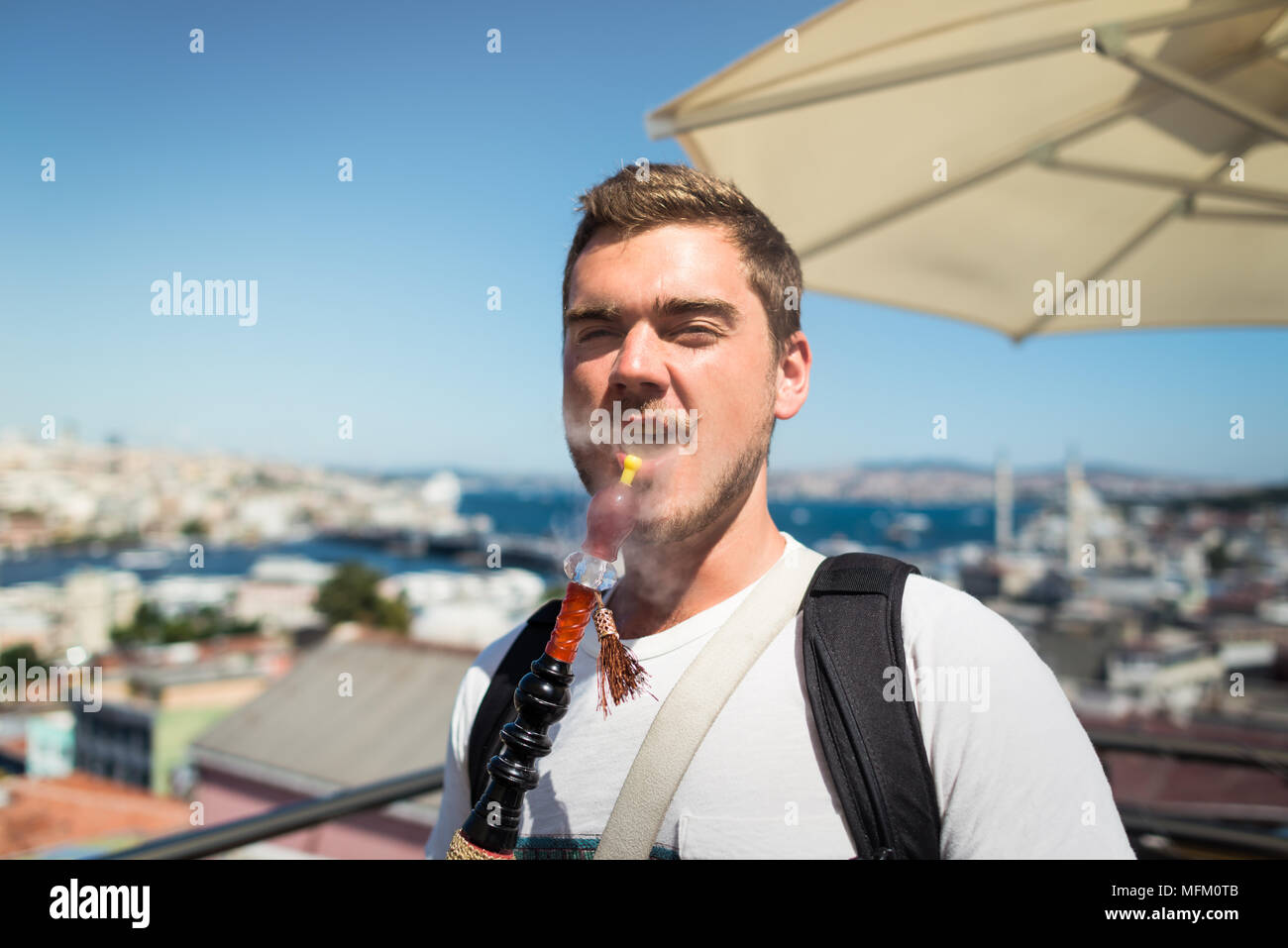 A young smiling man with mustache is smoking a hookah on the terrace overlooking the city Stock Photo