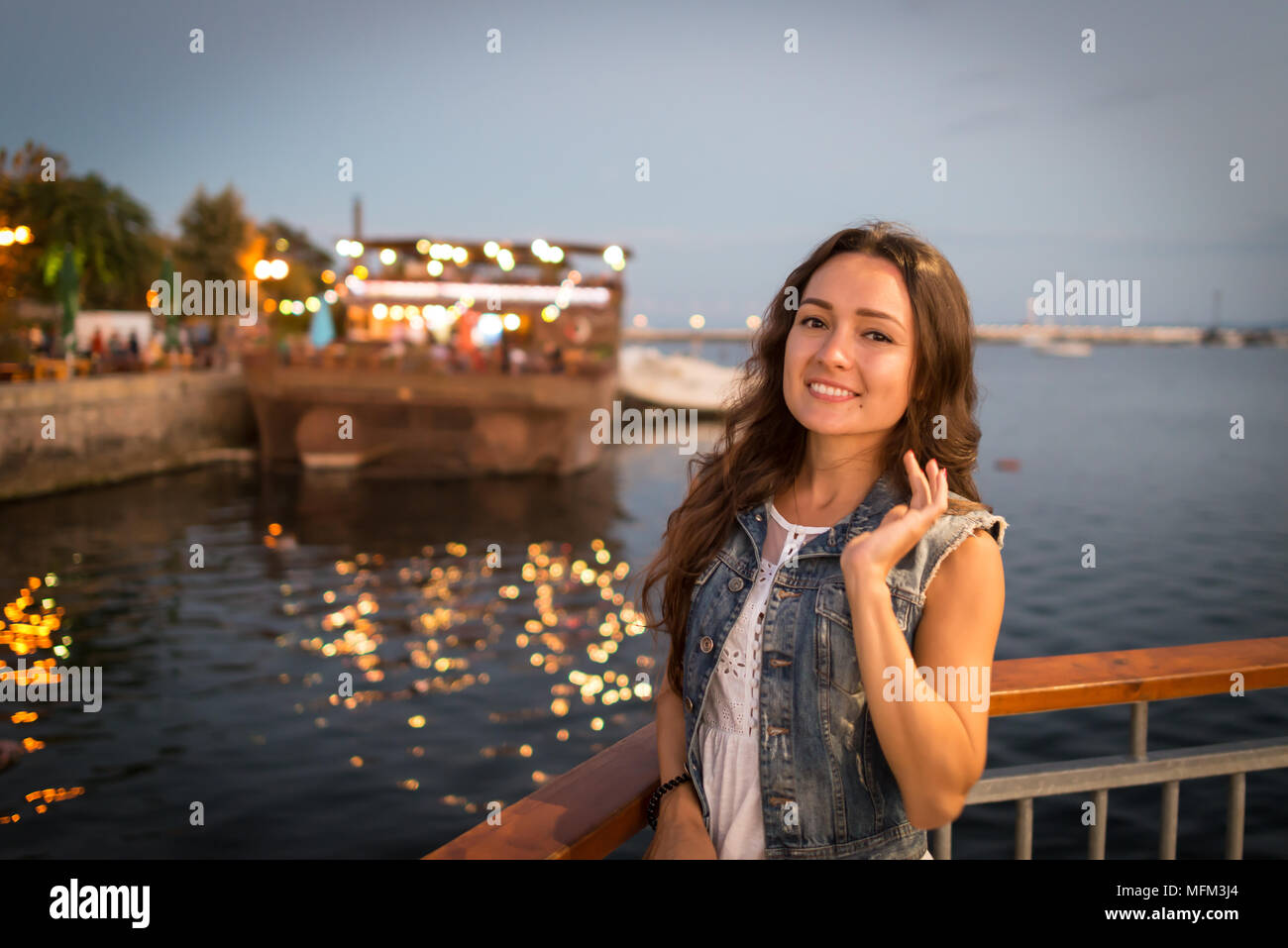 Pretty young woman walking on city promenade near sea in the evening. Soft colors. Street lights rays falls on her face. Happy girl enjoys vacation Stock Photo