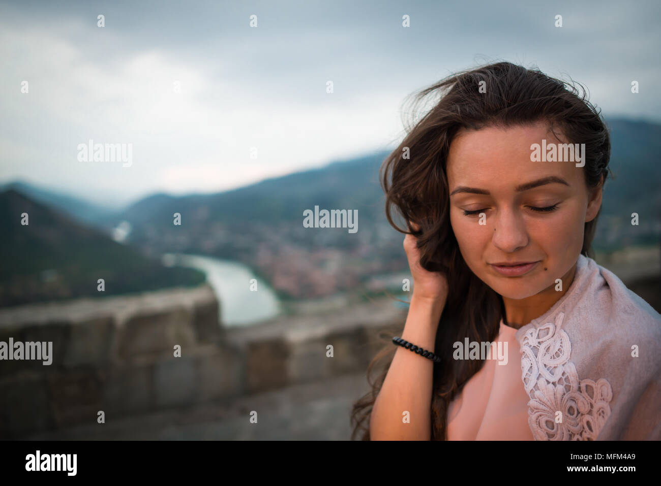 Young beautiful woman stands on background of mountains, river and small city downstairs. Pretty girl with closed eyes relaxes outdoor. Stock Photo