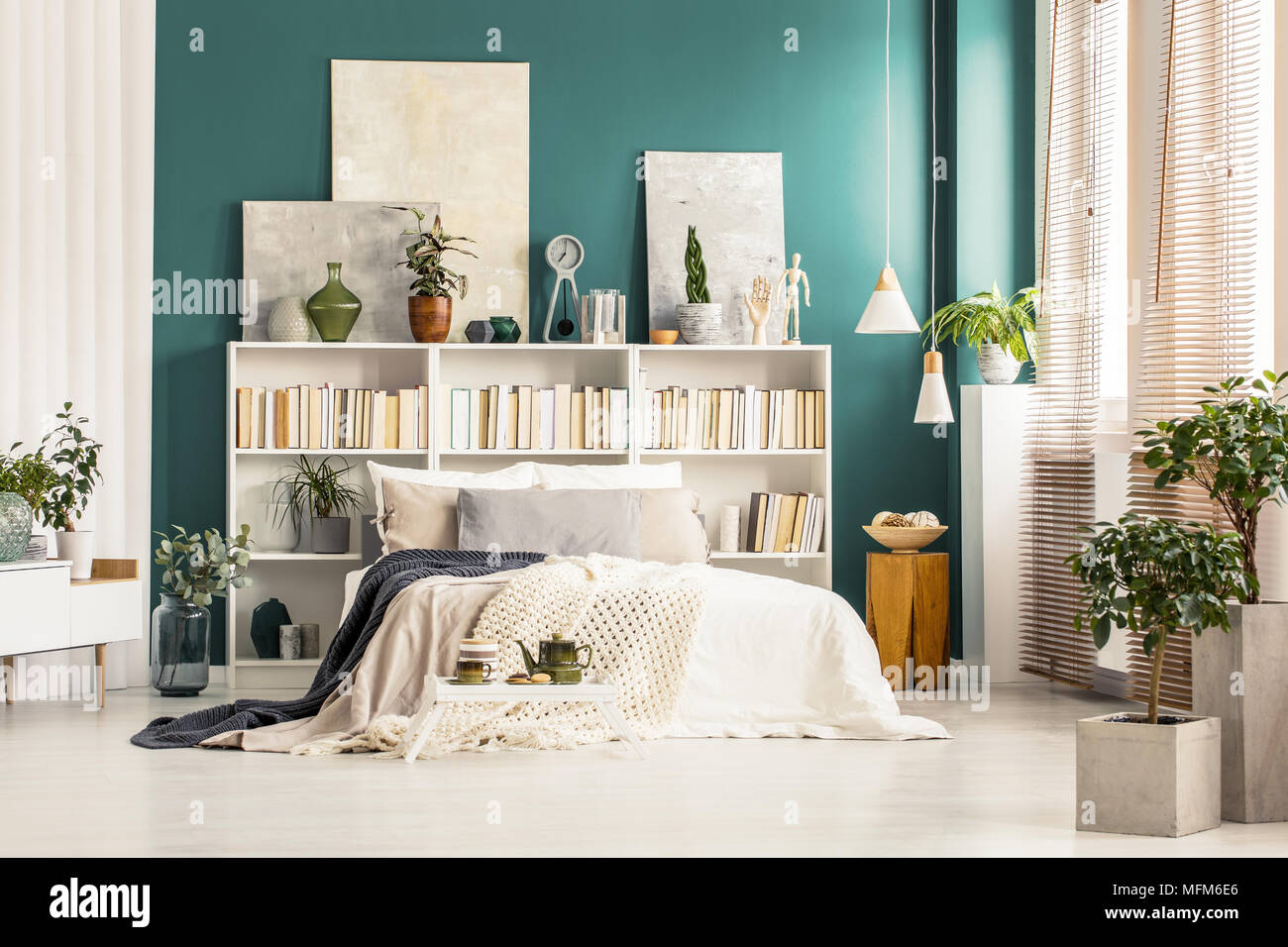 Bookcases with paintings on top standing against a dark green wall behind white bed in bright