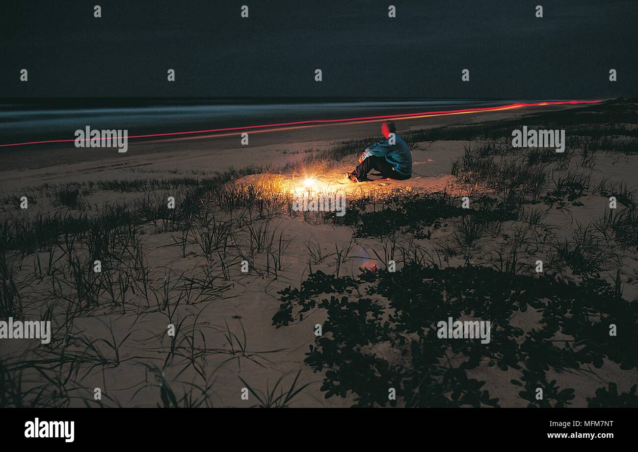 On the beach at night, Fraser Island, Queensland, Australia.  Light trails from departing 4WD vehicles illuminate the scene with a lone man sitting on Stock Photo