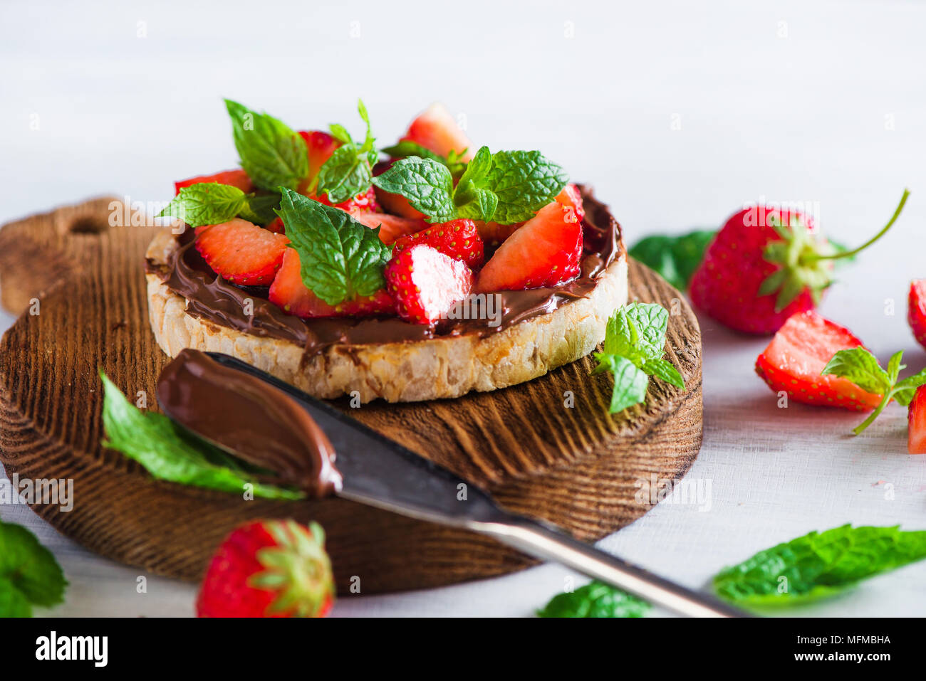 Crisp bread healthy snack with fresh strawberries, chocolate spread, mint leaves on a round cutting board with a dessert knife. Easy breakfast close-u Stock Photo