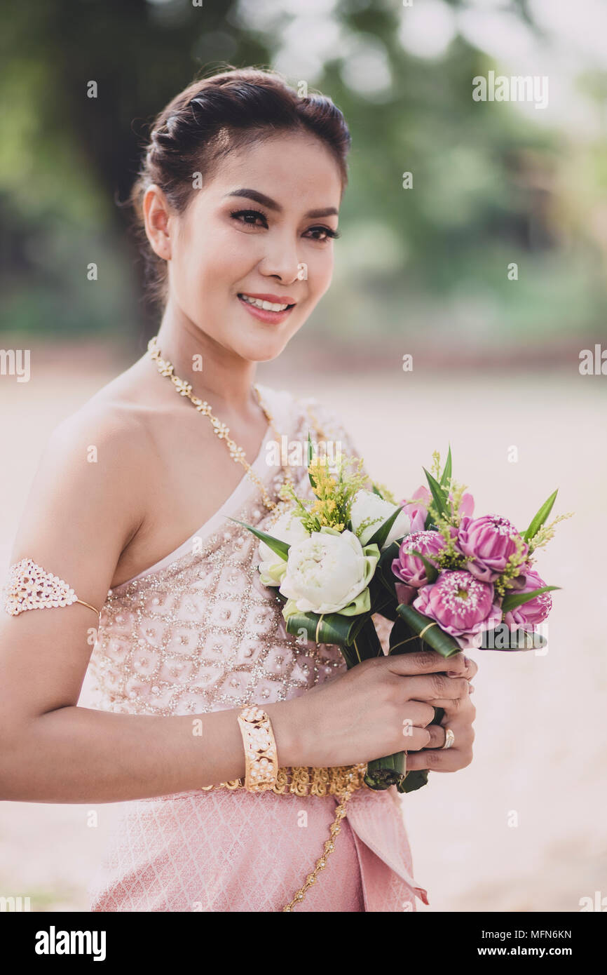 portrait of asian younger woman wearing thai tradition clothes holding pink lotus flower in hand toothy smiling face Stock Photo