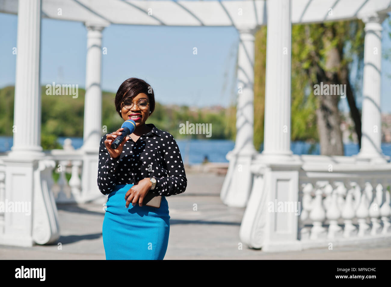 Stylish African American Model Girl Tv Presenter With Microphone In Glasses Blue Skirt And Black Blouse Posed Outdoor Against White Stone Arch With M Stylish African American Model Girl Tv Presenter With Microphone In Glasses Blue Skirt And Black Blouse Posed Outdoor Against White Stone Arch With M