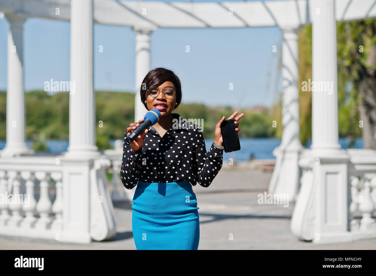 Stylish African American Model Girl Tv Presenter With Microphone In Glasses Blue Skirt And Black Blouse Posed Outdoor Against White Stone Arch With M Stylish African American Model Girl Tv Presenter With Microphone In Glasses Blue Skirt And Black Blouse Posed Outdoor Against White Stone Arch With M