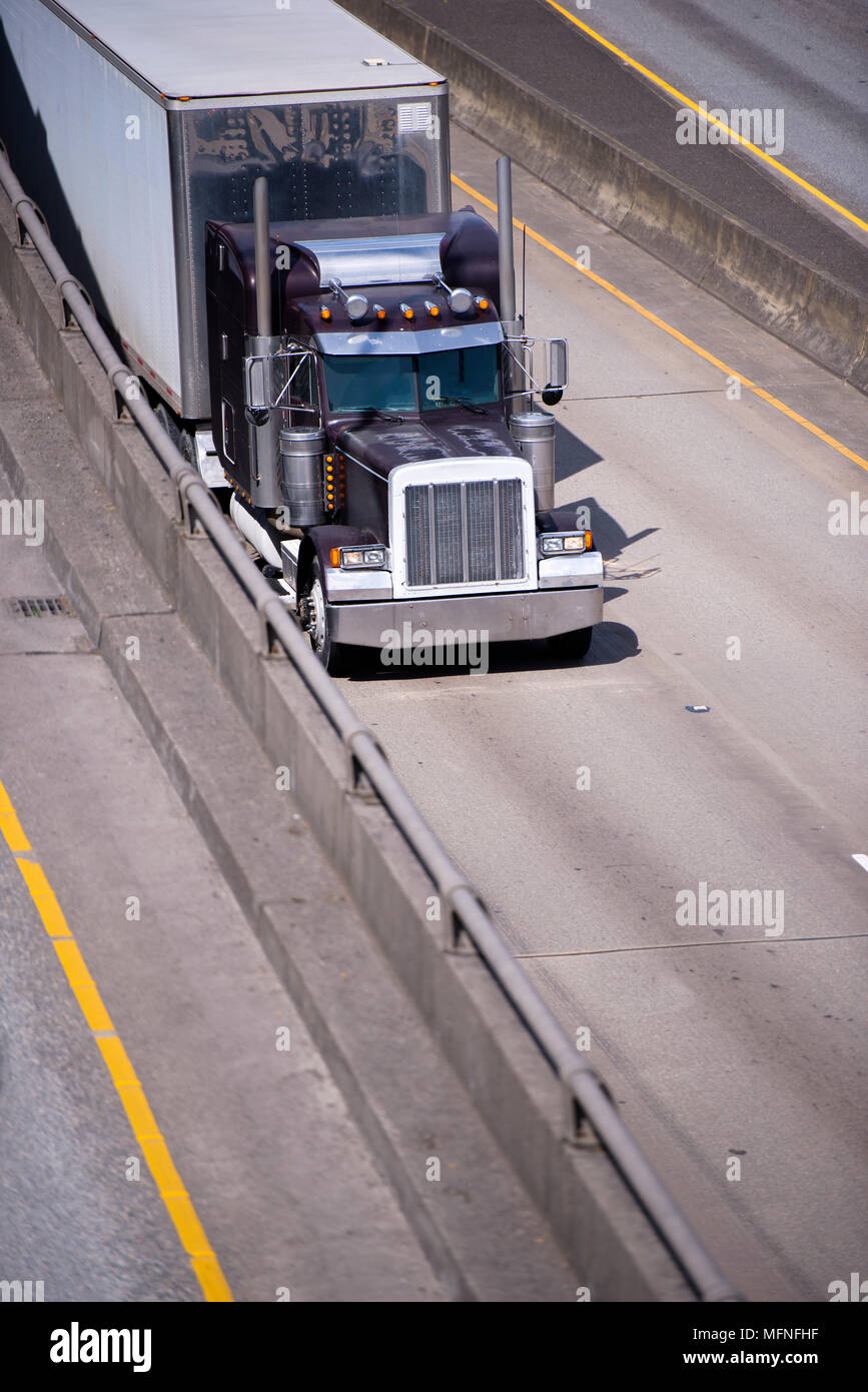 Dark old classic big rig semi truck idol with chrome accessories and tall exhaust pipes transporting cargo at dry van semi trailer with stainless stee Stock Photo