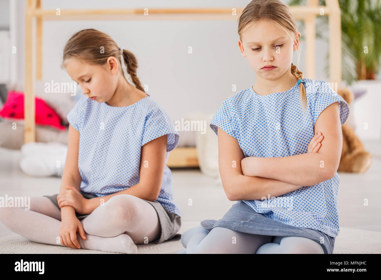 Twin sisters sad after a fight, looking down, sitting with her arms crossed in a bright kid's bedroom Stock Photo