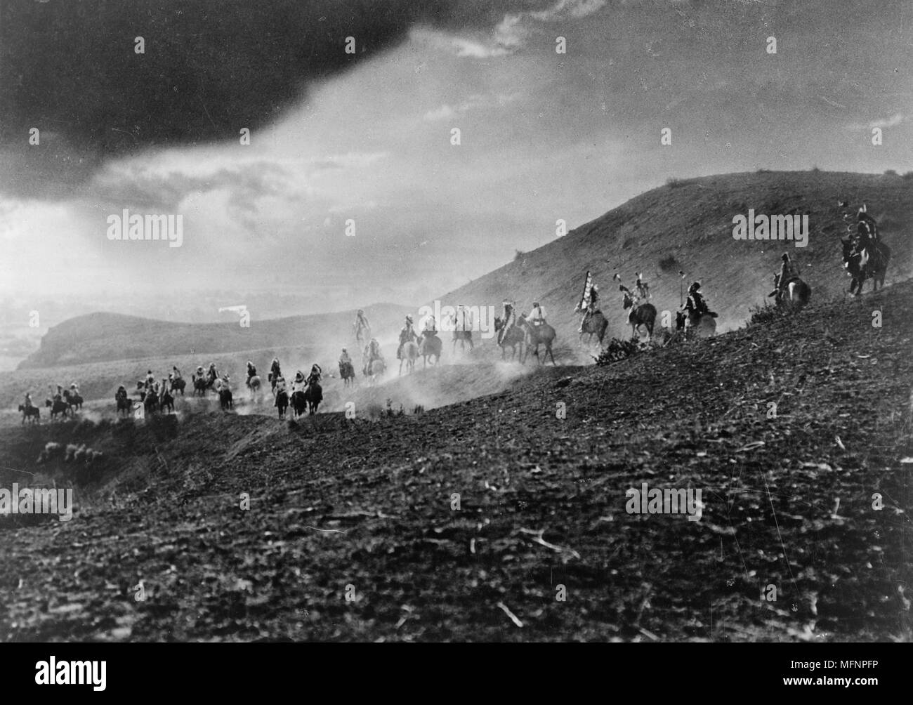 Group of Native Americans in war dress riding over hills in staged re-enactment, 1913. Photograph by by Joseph Kossuth Dixon Stock Photo