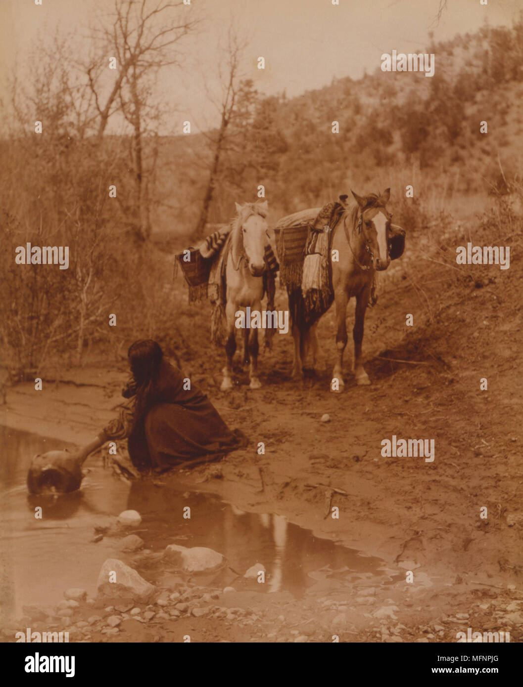 An Apache fills a container with water while two horses with panniers