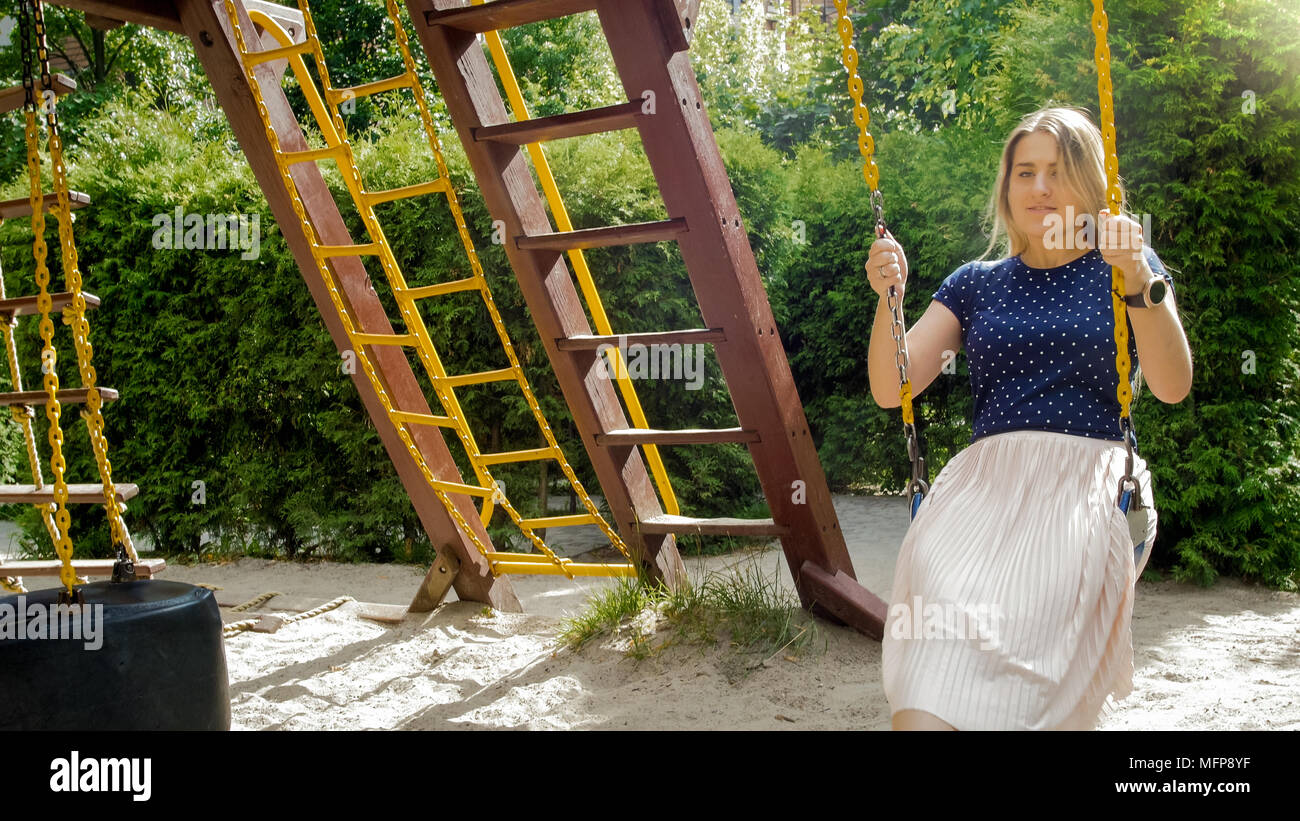 Portrait of smiling young woman sitting on swing at childs playground ...