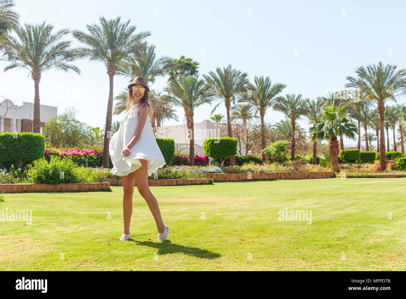 Woman on a holiday with sunglasses and white dress relaxing outdoor on a beautiful green park with palm trees. Stock Photo