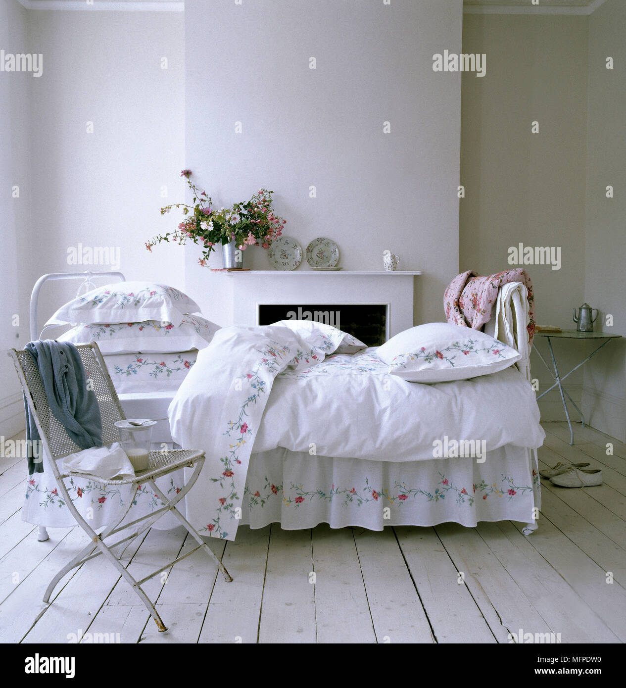 Neutral country bedroom with painted wood floor, wrought iron bed, floral linen, fireplace, and metal folding chair. Stock Photo