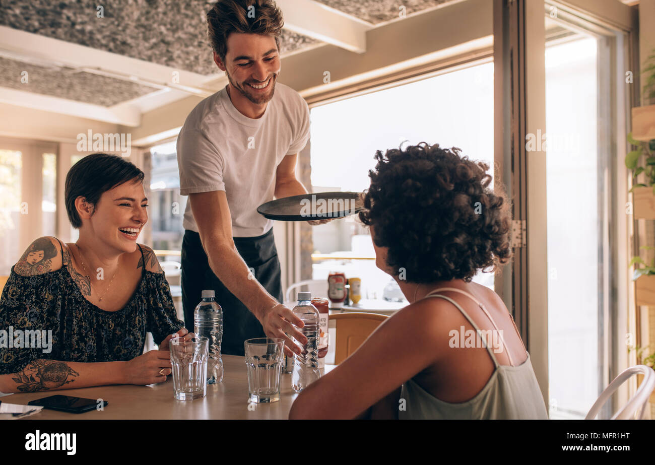 Smiling female friends at a cafe with waiter serving water. Two young women sitting at a restaurant. Stock Photo