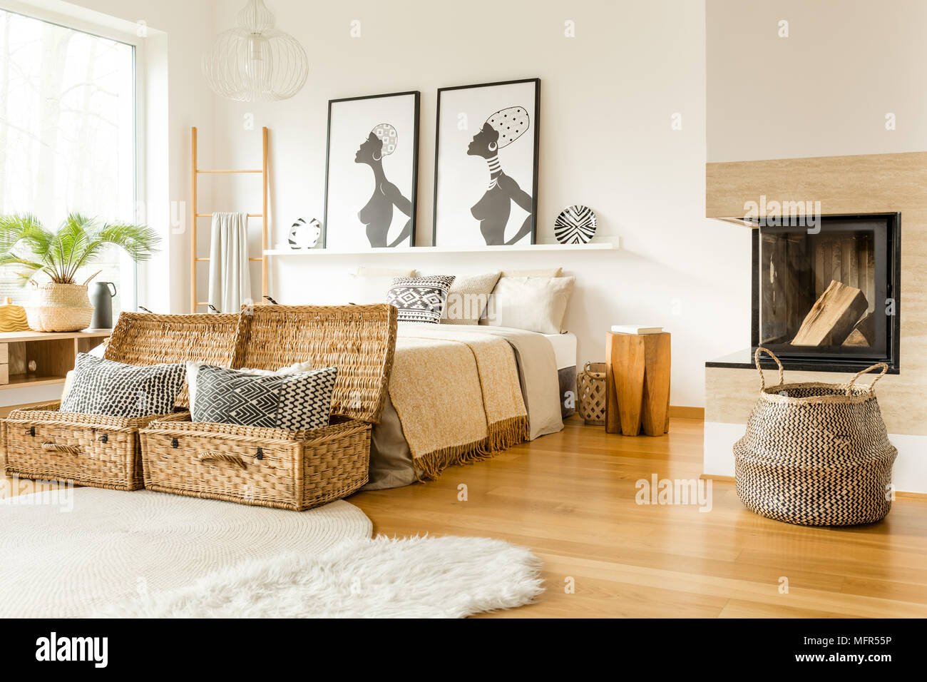 Fireplace, wattle boxes with pillows, bed and African posters in a boho bedroom interior Stock Photo