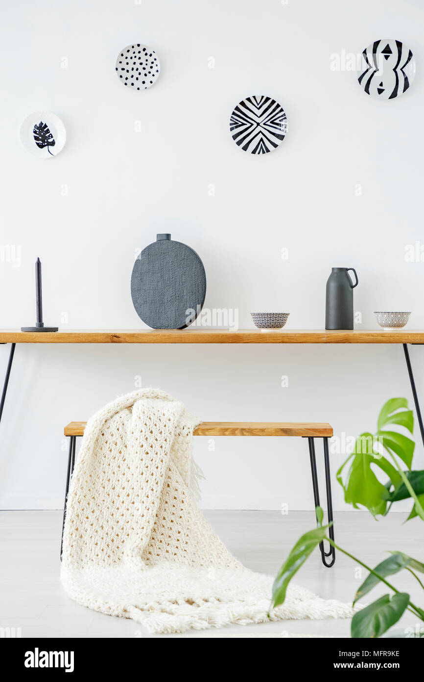 Knit blanket on a bench at wooden table with grey vase against white wall with plates in flat interior Stock Photo