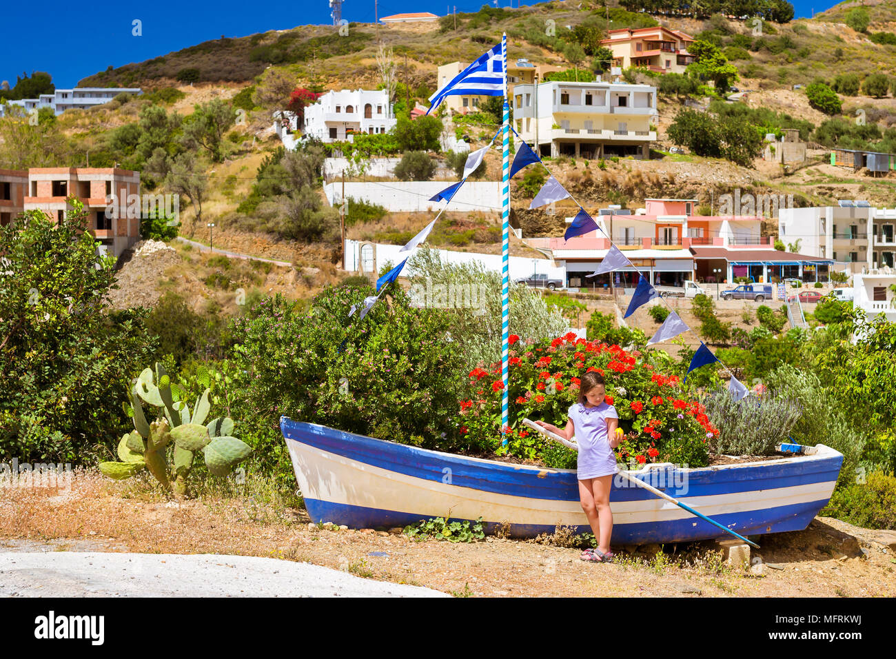 Girl with yellow lemon in hand posing against old fishing boat with Greek flag, stands on shore and decorated by flower bed. Tourist attraction Varkot Stock Photo
