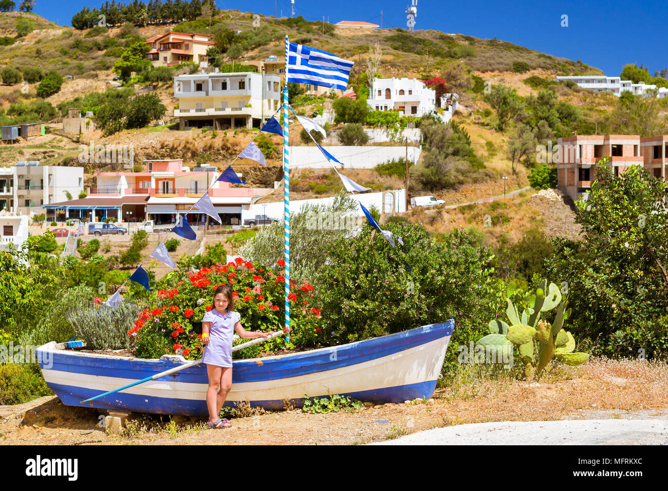Girl with yellow lemon in hand posing against old fishing boat with Greek flag, stands on shore and decorated by flower bed. Tourist attraction Varkot Stock Photo