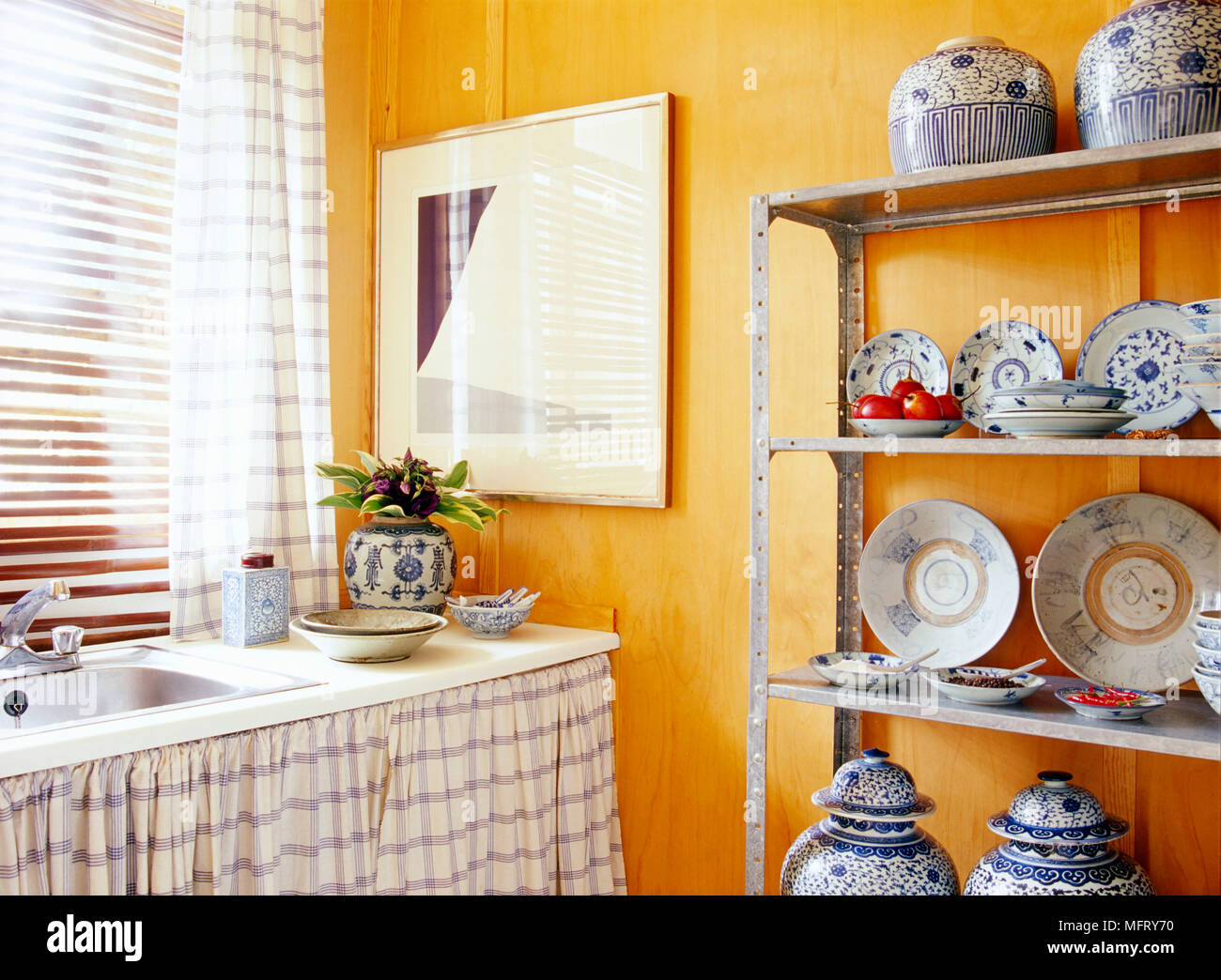 A detail of a modern yellow kitchen showing a sink unit with curtain metal shelving unit with a display of oriental ceramics; Stock Photo