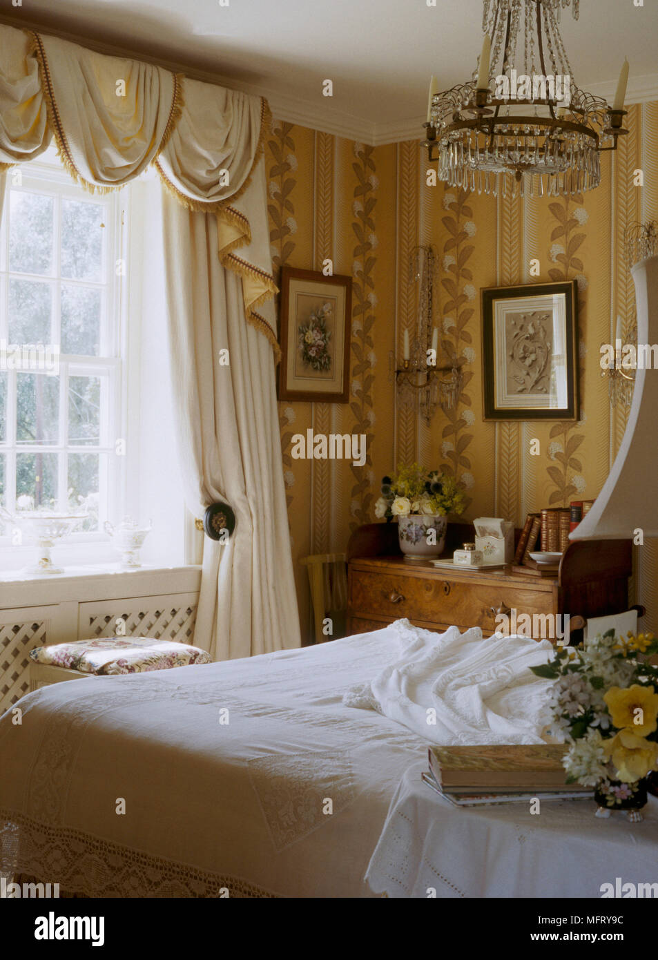 A detail of a traditional yellow bedroom with pattern wallpaper swag curtains double bed glass chandelier Stock Photo