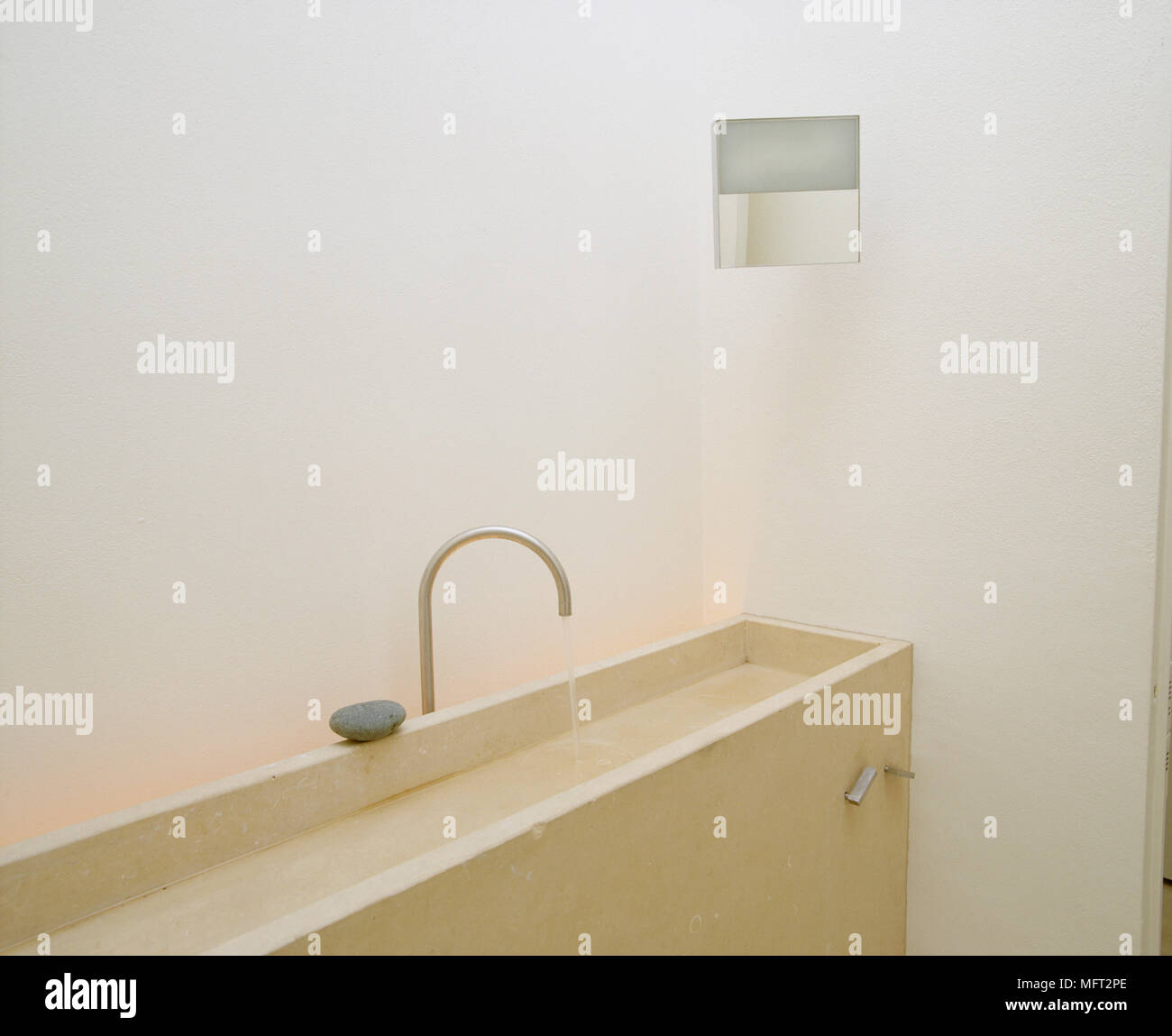 A detail of a modern, neutral, minimalist bathroom showing a stone trough sink with water running from tap Stock Photo