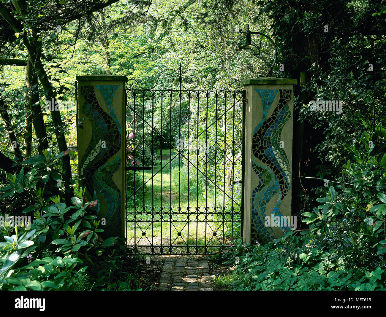 Wrought iron gates with pillars decorated with mosaic tiles in a shady garden, Stock Photo