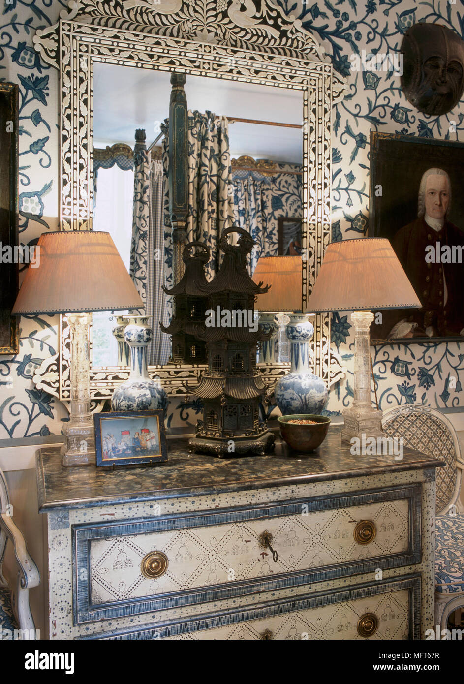 A detail of a traditional, blue bedroom with pattern wallpaper, decorative painted chest of drawers, ornate mirror, pair of lamps Stock Photo
