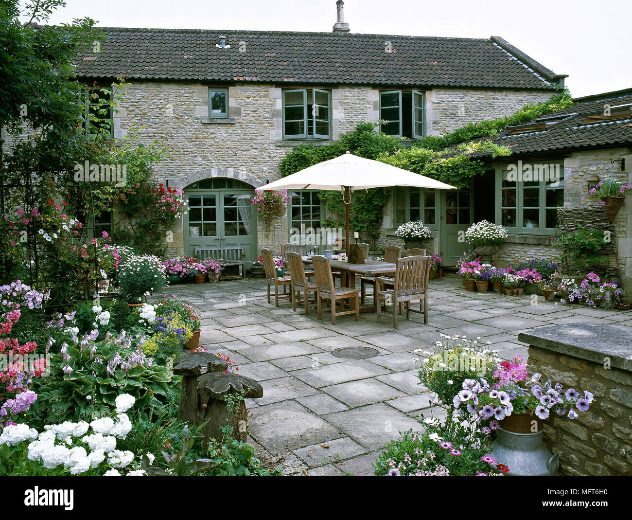 Exterior of the back terrace of a stone country house with tile floor, potted flowers, and an outdoor dining table and chairs. Stock Photo
