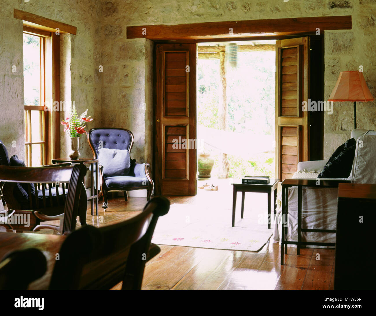 Rustic sitting room with stone walls wood floor a couch and wooden armchairs and an open doorway out to a back patio. Stock Photo