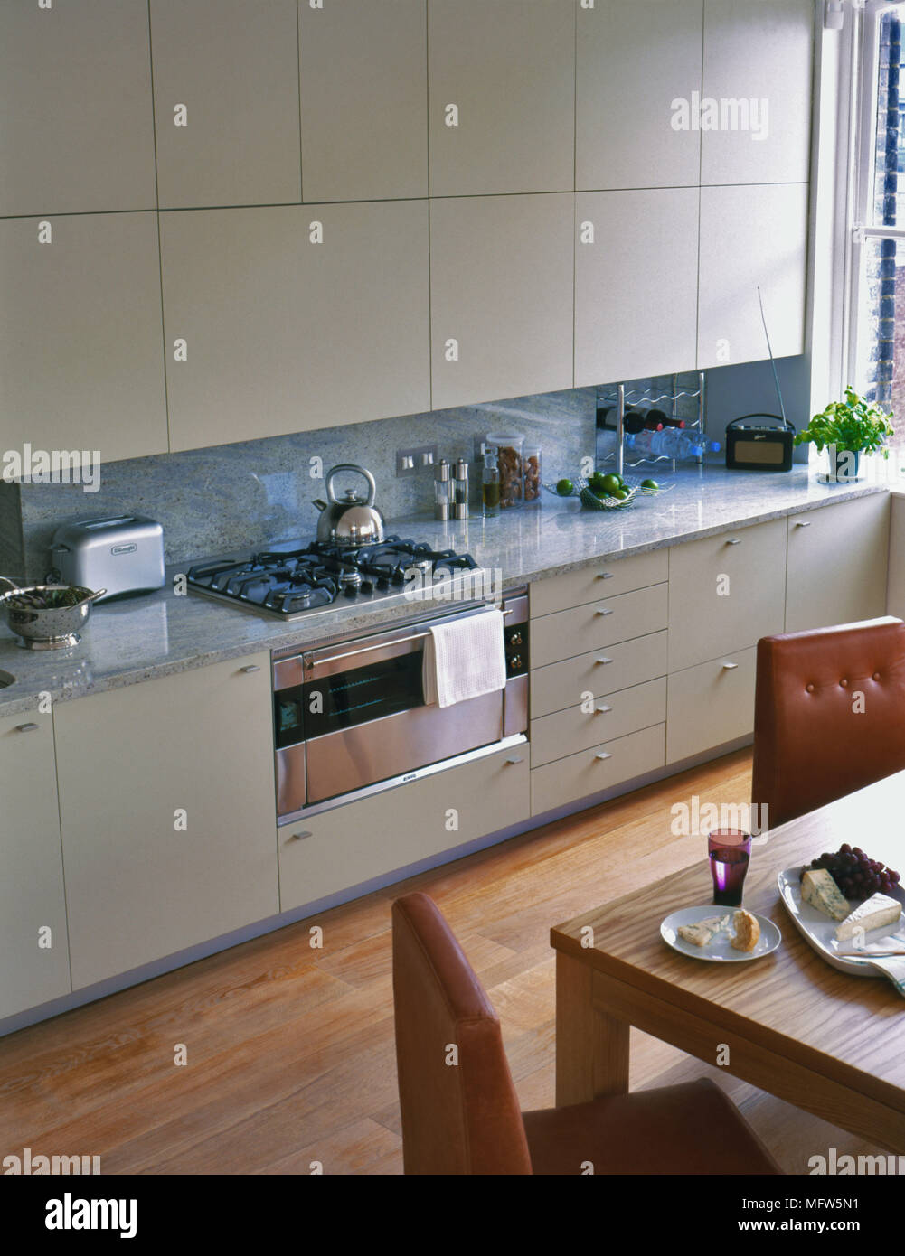 A modern kitchen with minimalist kitchen units above a stainless steel oven with a wooden kitchen table. Stock Photo