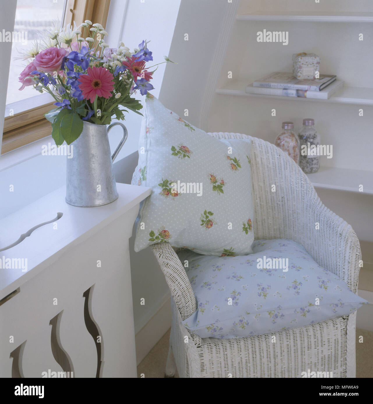 A detail of a country bedroom showing a Lloyd Loom wicker armchair with floral cushions flower arrangement in a galvanised jug Stock Photo