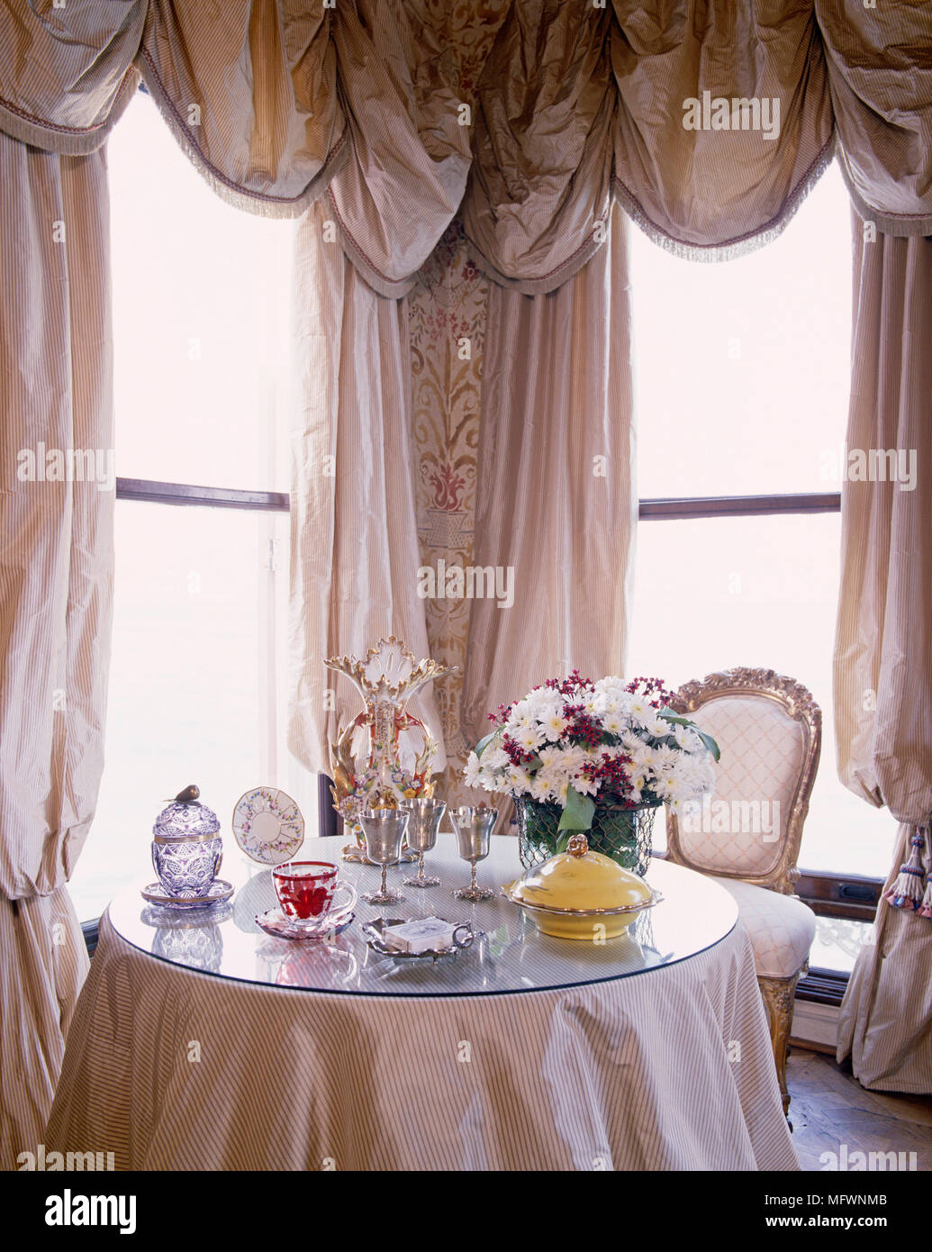 Round table with glass top in bay window dressed with swag curtains Stock Photo