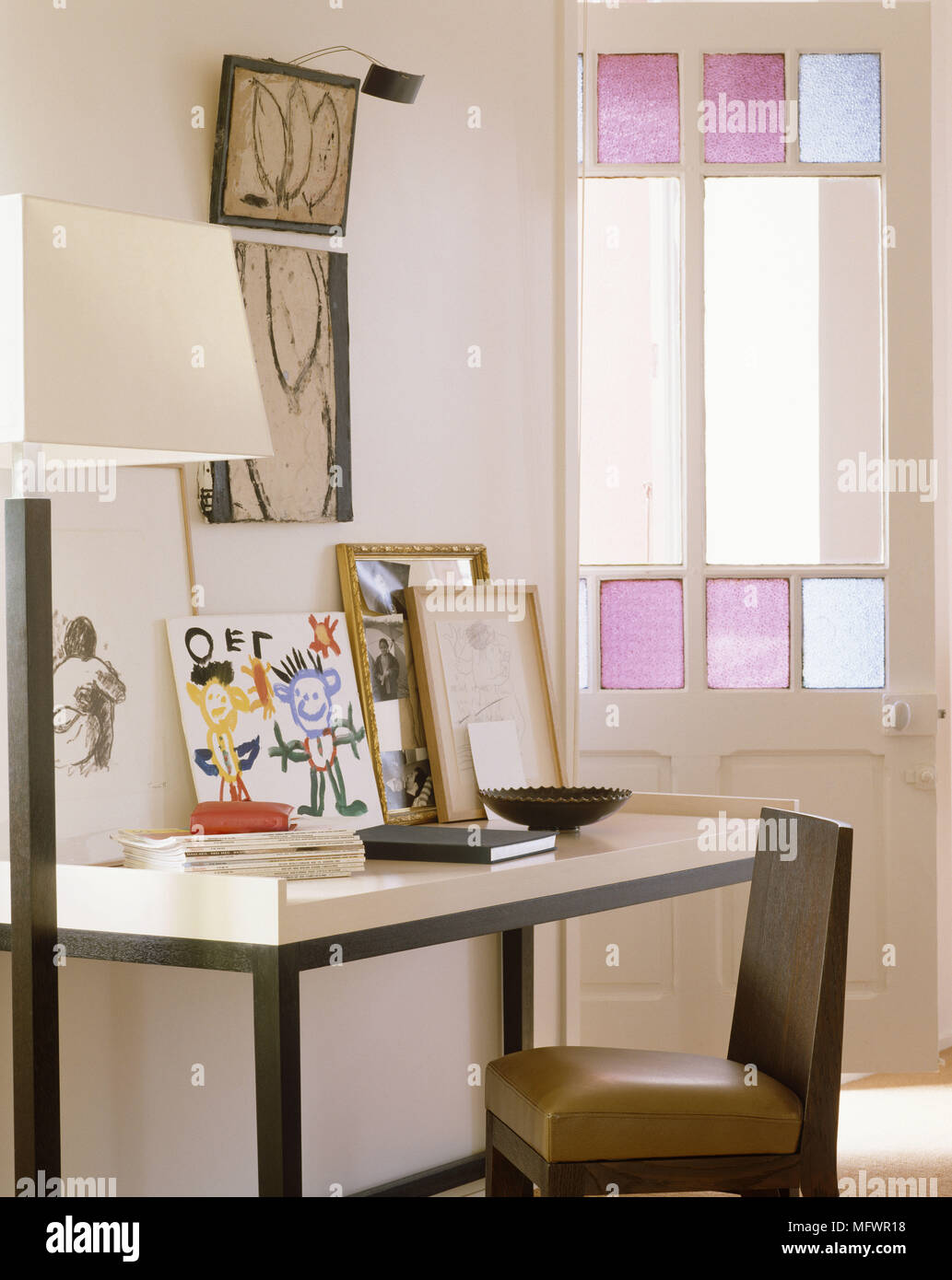 Various artwork above writing desk with upholstered chair next to door with stained glass Stock Photo