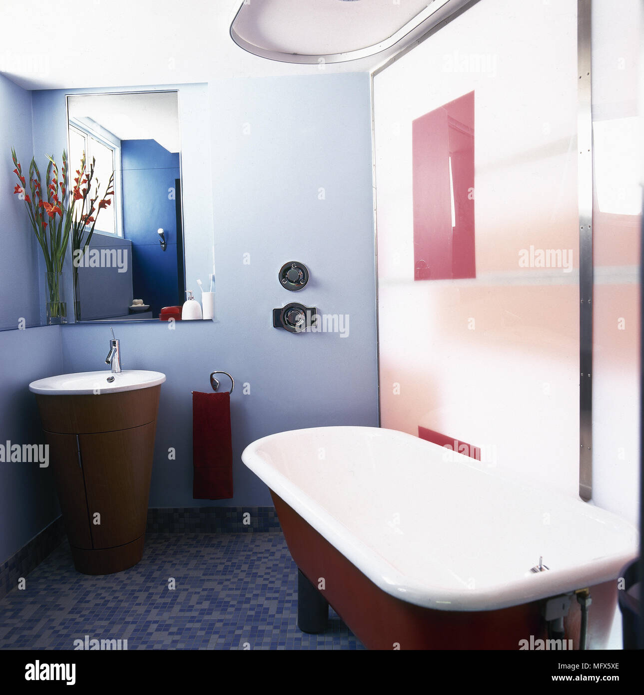 Modern, tiled bathroom with a roll top bathtub, blue accent wall, and a washbasin in a cylindrical cabinet. Stock Photo
