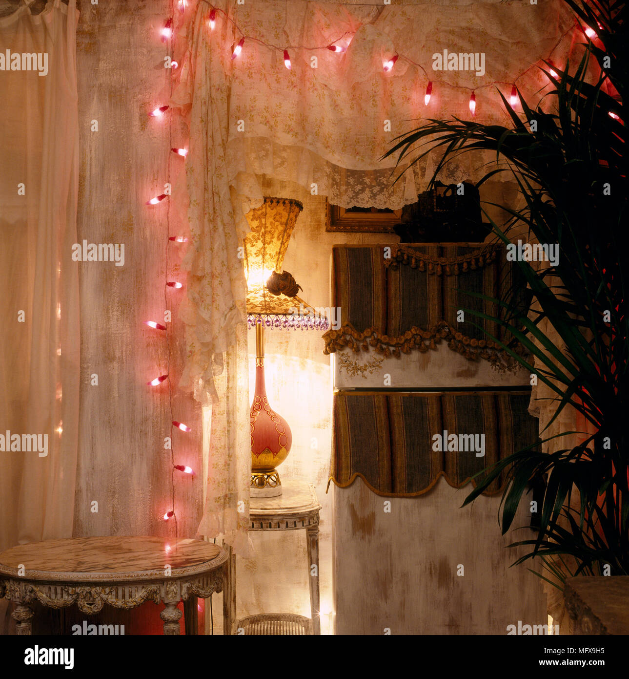 A detail of a romantic bedroom with lit fairy lights draped around lace curtains, lit lamp giving a subtle, low light ambient Stock Photo