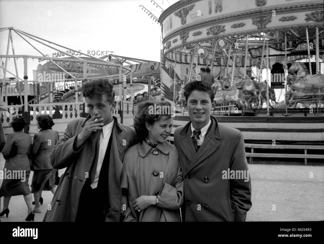 3 well dressed fashionable teenagers, 2 boys and a girl, on a cold day at a British funfair in the 1950s, one is smoking a cigarette. Stock Photo