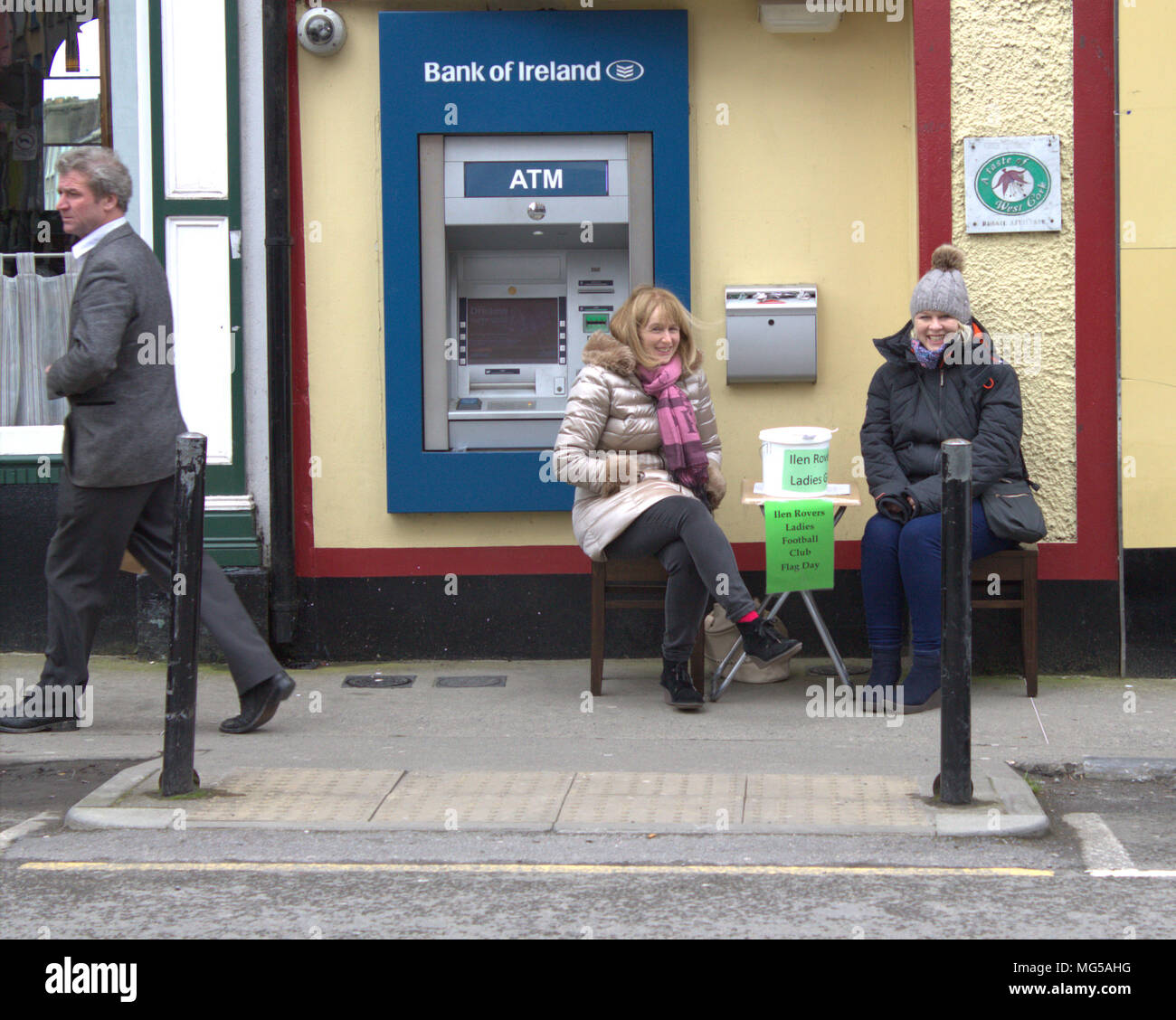 2 mid adult females wearing heavy coats trying to keep out the cold wind, are sat next to an ATM with a collection bucket as a single male walks by. Stock Photo