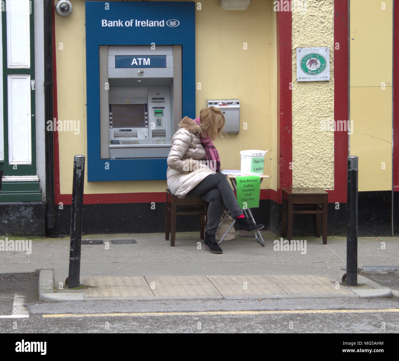single mid adult female wearing a heavy coat trying to keep out the cold wind, sitting on the sidewalk next to an ATM with a collection bucket. Stock Photo