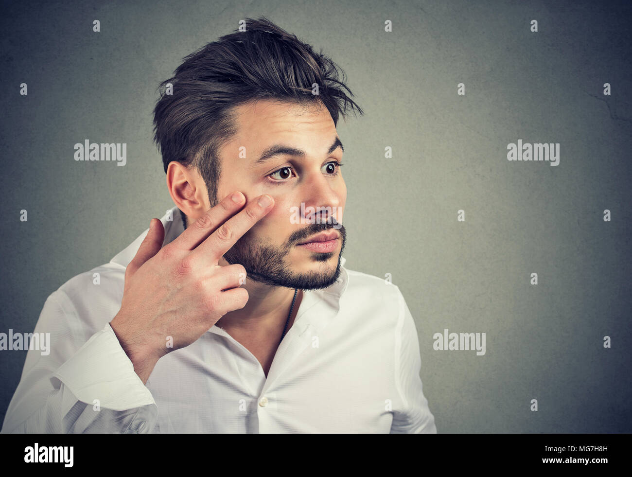 Young man pulling down eyelid checking his eye looking in mirrow feels unwell has black circles Stock Photo