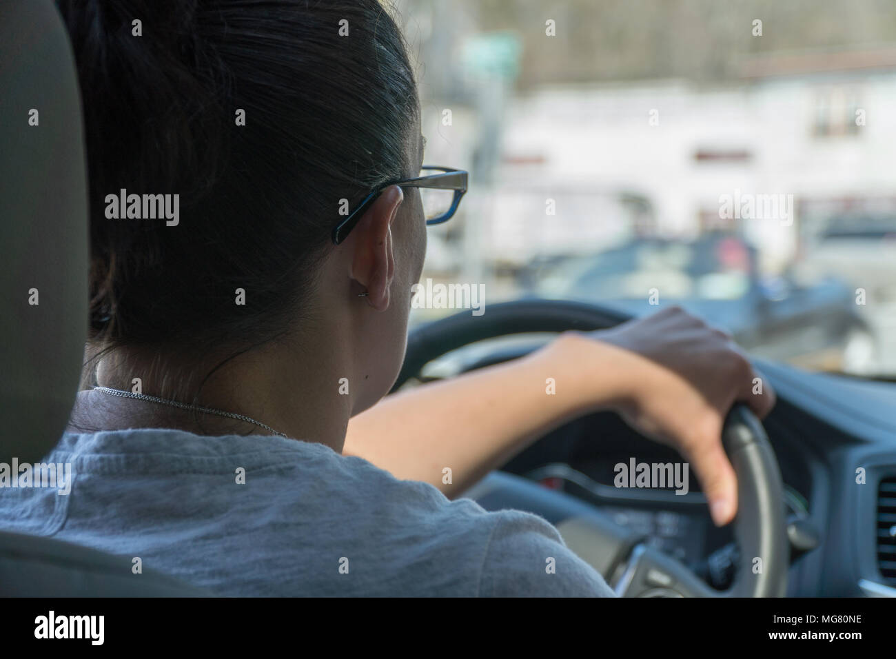 Over the shoulder POV young woman driving a car on a busy urban street road during the day time. Stock Photo