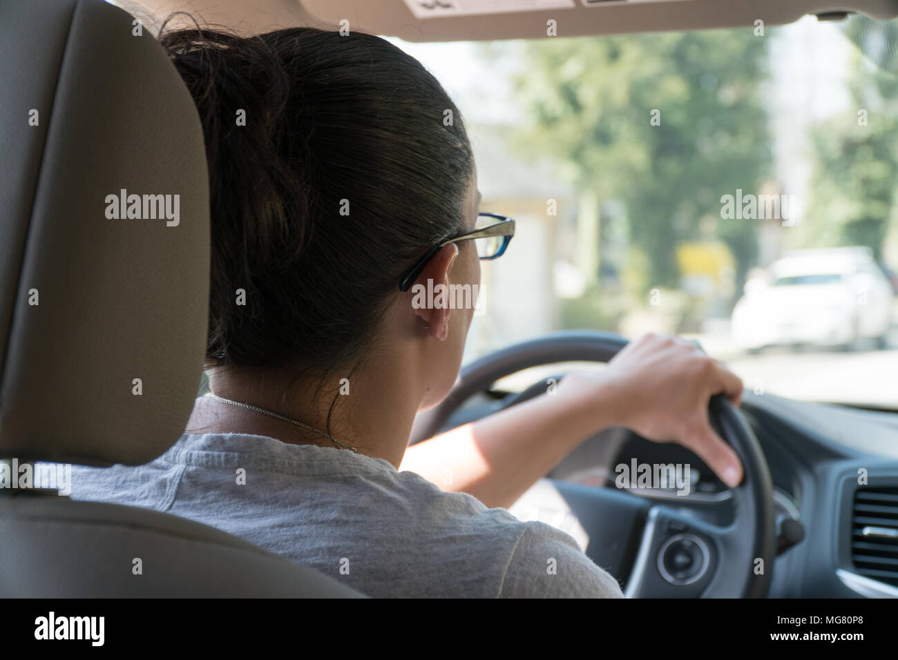 Over the shoulder POV young woman driving a car on a suburban street past houses and parks along road during the day time. Stock Photo