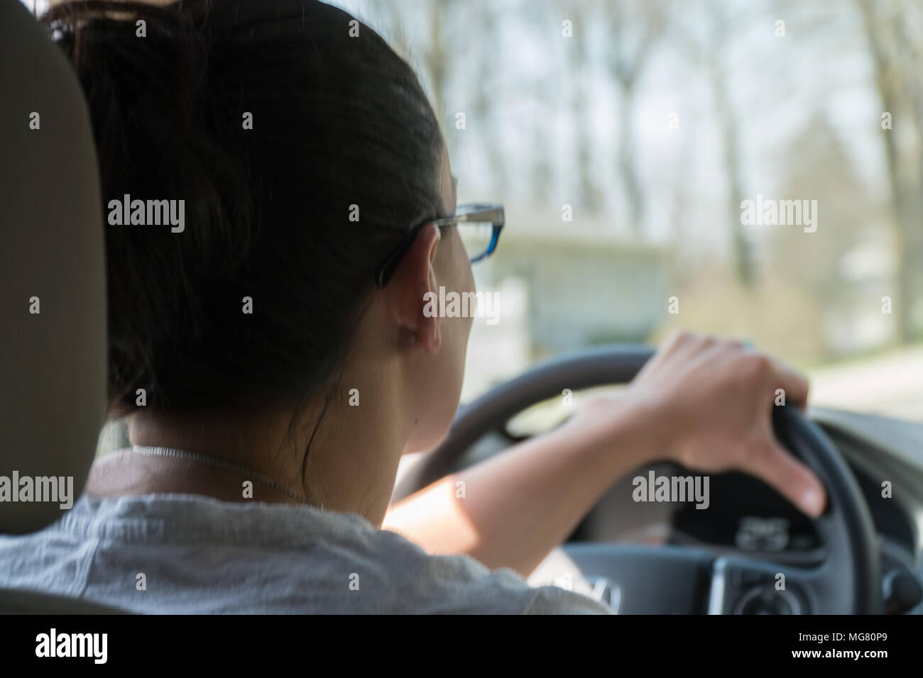 Over the shoulder POV teen girl driving a car on a rural road road during the day time. OTS. Stock Photo