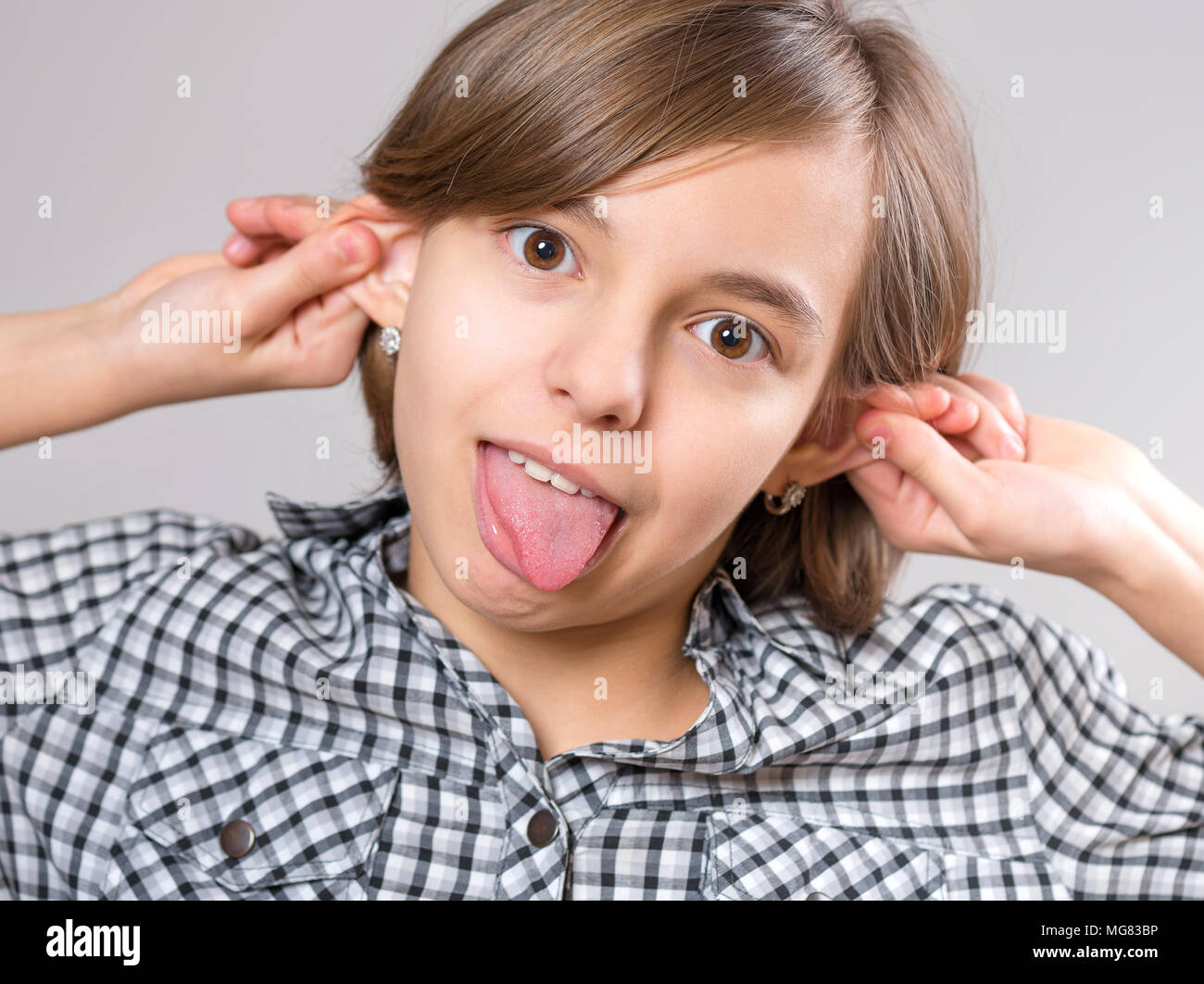 Silly girl making grimace - funny monkey face. Child with big ears, on gray background. Emotional portrait of caucasian teenager looking at camera. Stock Photo