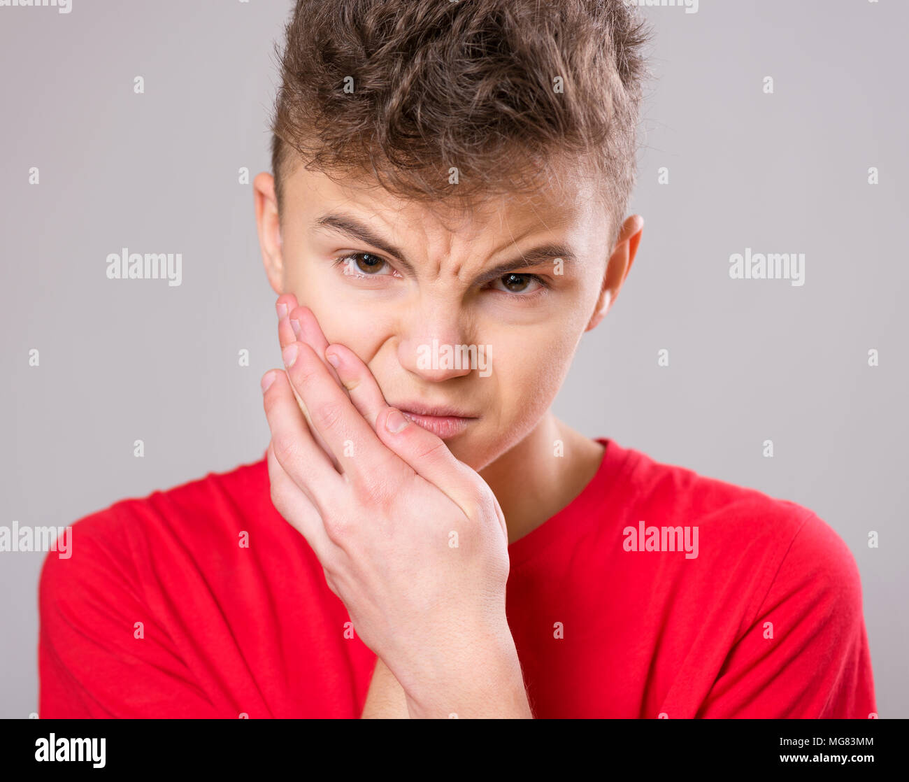 Casual guy have toothache, on gray background. Emotional portrait of teen boy wearing red t-shirt. Sad child with tooth pain. Dental problem - teenage Stock Photo