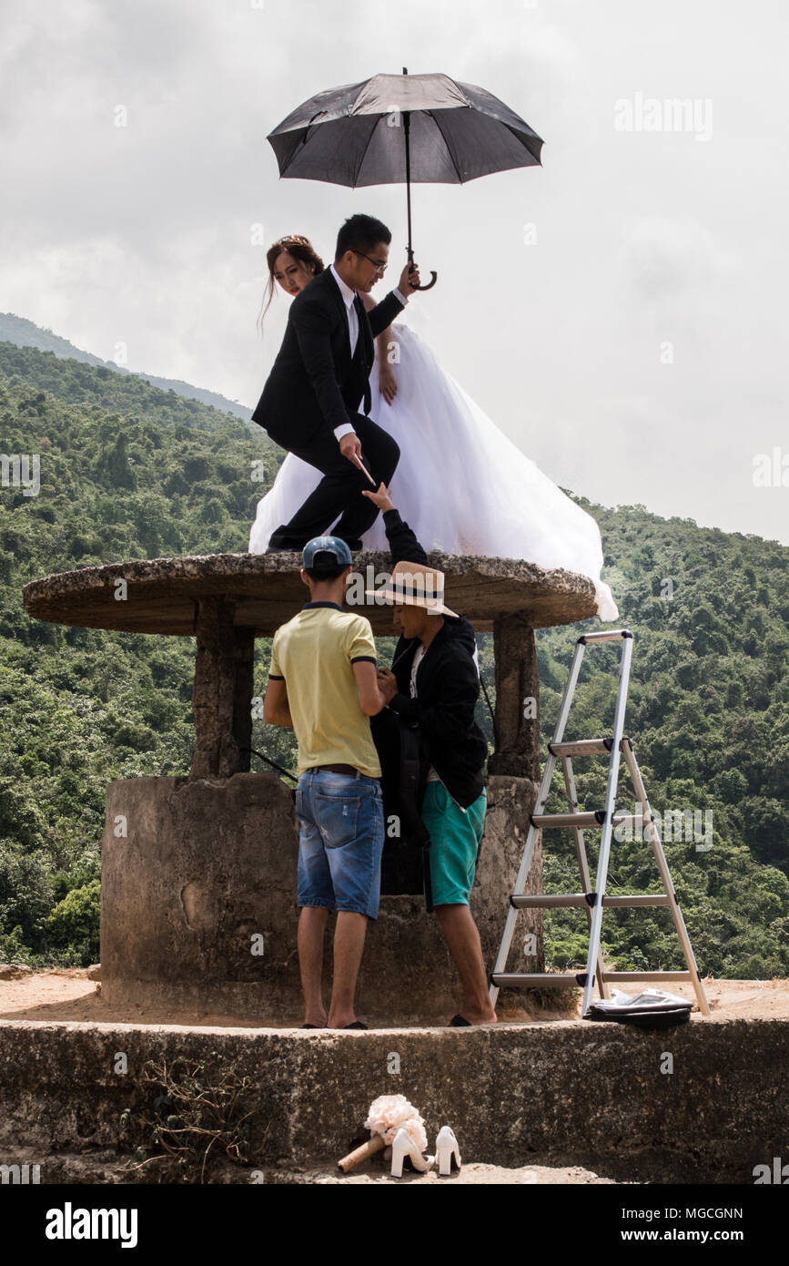 Newly-wed couple, posing for photographs, perched on an old well, holding umbrella, while photographer and assistant talk below, Hue, Vietnam Stock Photo