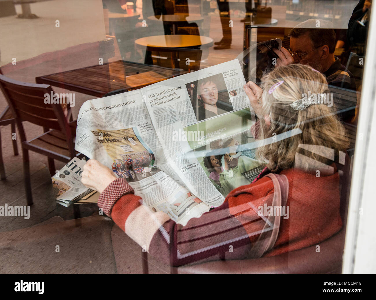 Woman sitting in in cafe, reading newspaper, seen through glass window, reflection of outdoor furniture on window Stock Photo