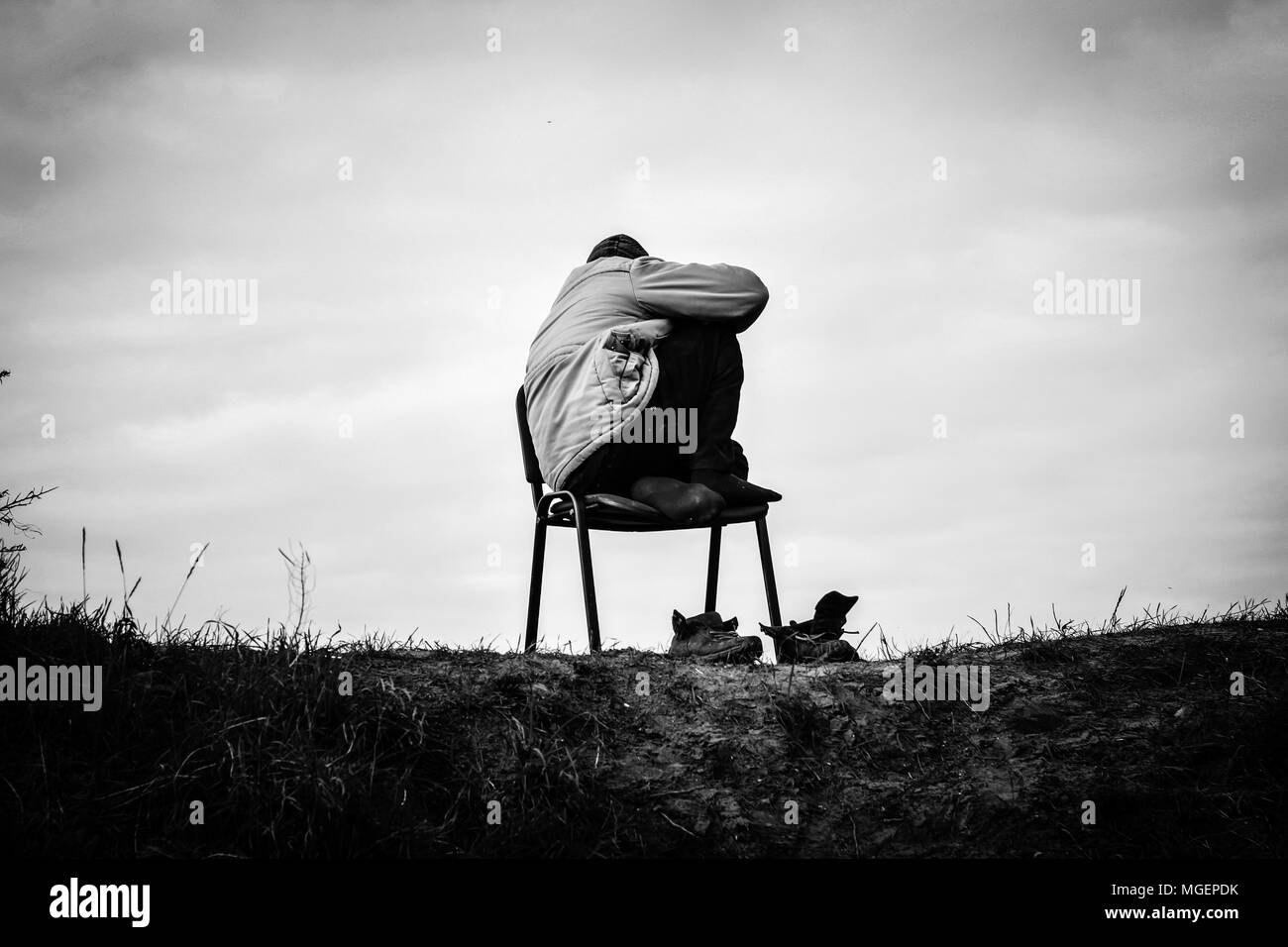 A poor refugee refugee in the Jungle field of Calais crouches hooded on a chair at the top of the hill taking off his shoes Stock Photo