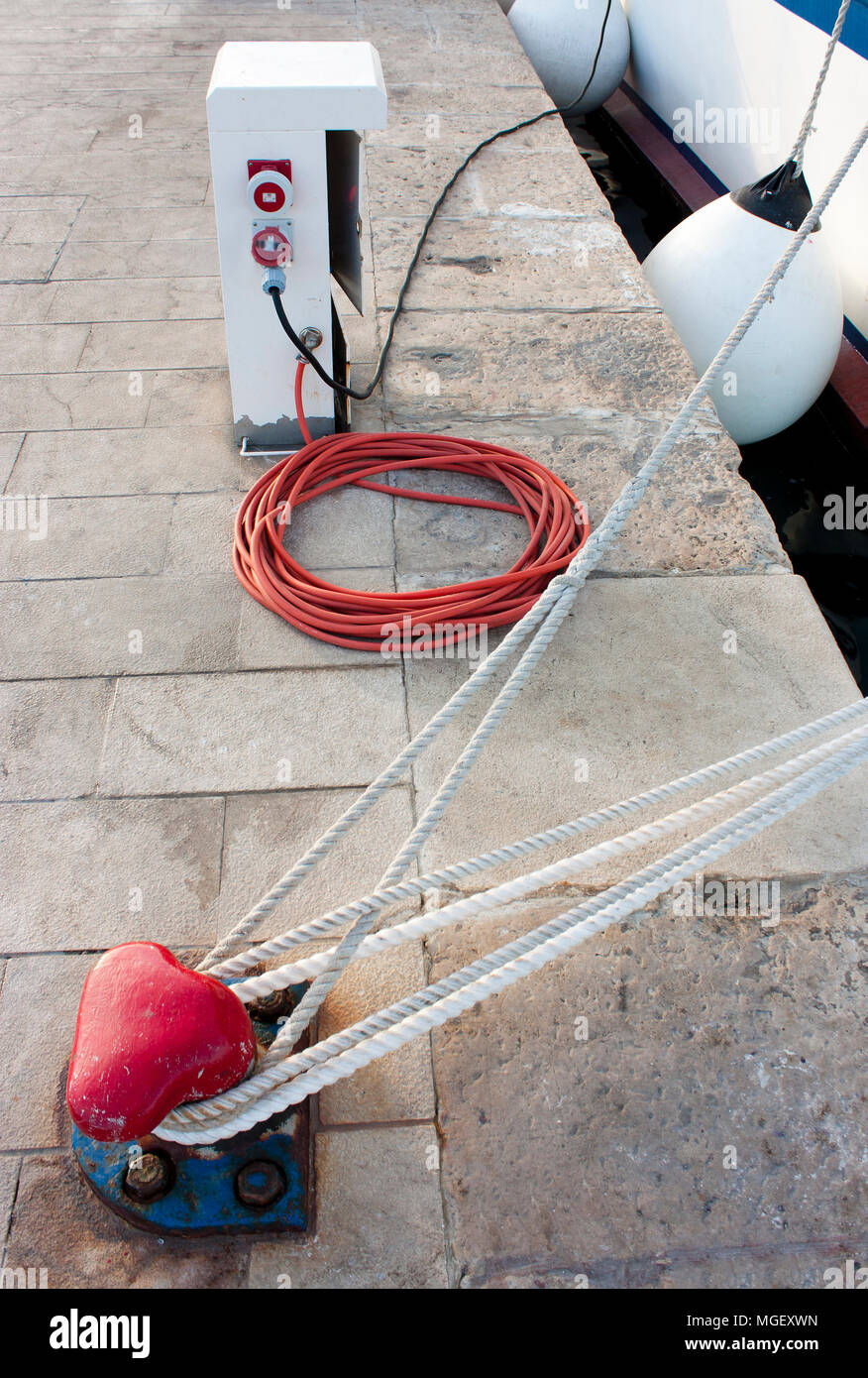 Electrical and water supply pedestal with hose and cable , bitt and a rope on the dock fastened to a boat Stock Photo