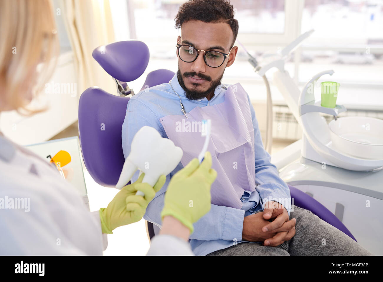 Unrecognizable blond-haired dentist explaining mixed-race young patient how to brush teeth correctly while having appointment at modern office Stock Photo