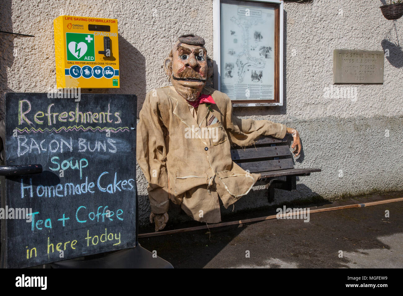 Old man scarecrow seated on seat, next to emergency defibrillator box in Wray, Lancaster, UK. 28/04/2018. Wray Scarecrow Festival and village cafe.The villagers  are back again to creating some weird, wacky and wonderful scarecrow creations to surprise and delight visitor. Each year visitors wander around Wray village, discovering the host of scarecrows that have popped up in village gardens. Credit: MediaWorldImages/AlamyLiveNews Stock Photo
