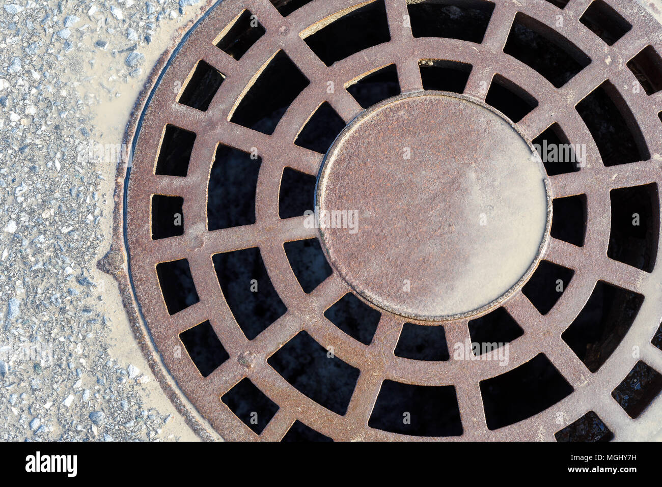 Cast iron metal rain manhole cover in tarmac with fresh mud stained rain water surrounding the entrance grills of this heavy entrance lid Stock Photo